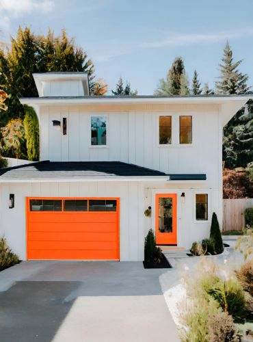 A modern two-story house with white siding and a black roof. The garage door and front door are bright orange. Lush greenery surrounds the home, with tall trees in the background and neatly arranged shrubs in the foreground.