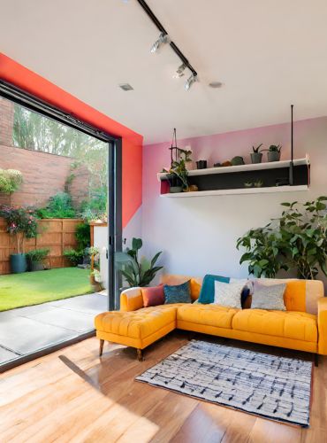 A modern living room featuring a yellow sectional sofa with colorful pillows. A patterned rug is on the wooden floor. To the left, large glass doors open to a green outdoor space with plants. A shelf above holds small potted plants against a pink accent wall.