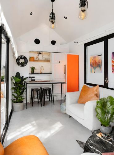 An interior view of a modern tiny home featuring a small dining table with black stools, a white couch with an orange pillow, and an orange wall. Green plants are placed in pots, and artwork is displayed on the walls, creating a bright, stylish space.