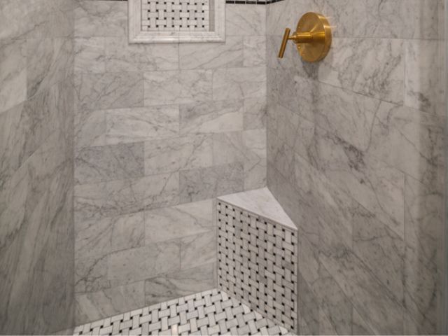A modern shower featuring marble walls with light and dark gray veining. The shower has a built-in bench covered with small black and white tiles. A gold shower handle is mounted on the wall, and a small square niche is visible for toiletries.